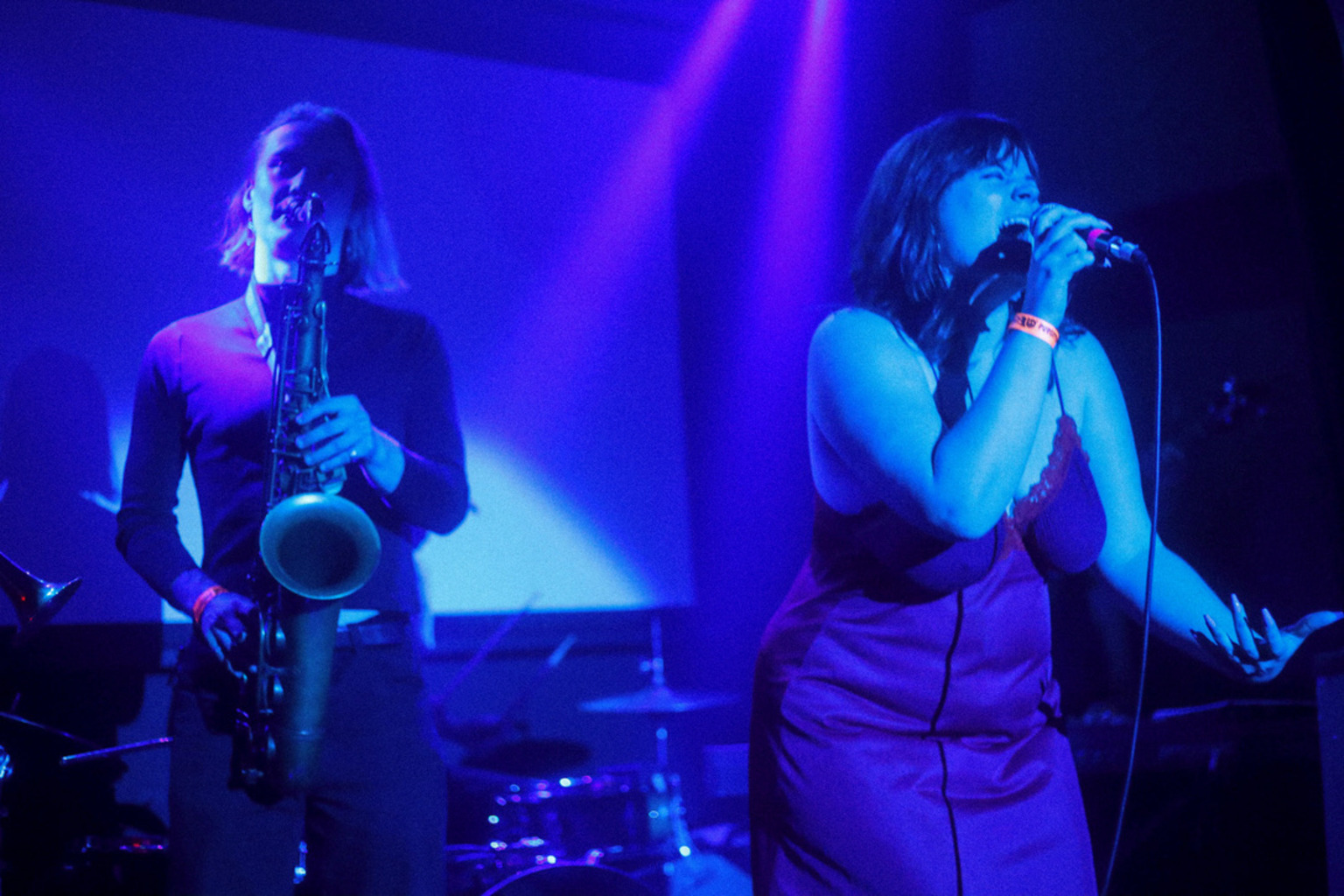 Madeline Moneypenny sings at the front of a moodily lit stage (right), backed by a saxophonist (left).