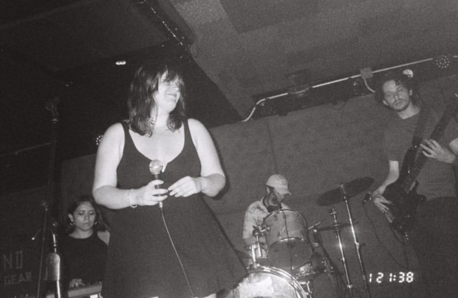 A black and white photograph from an old digital camera. Madeline dances on stage with the band at a show. There's a digital watermark in the bottom-right corner. It reads 121:38.