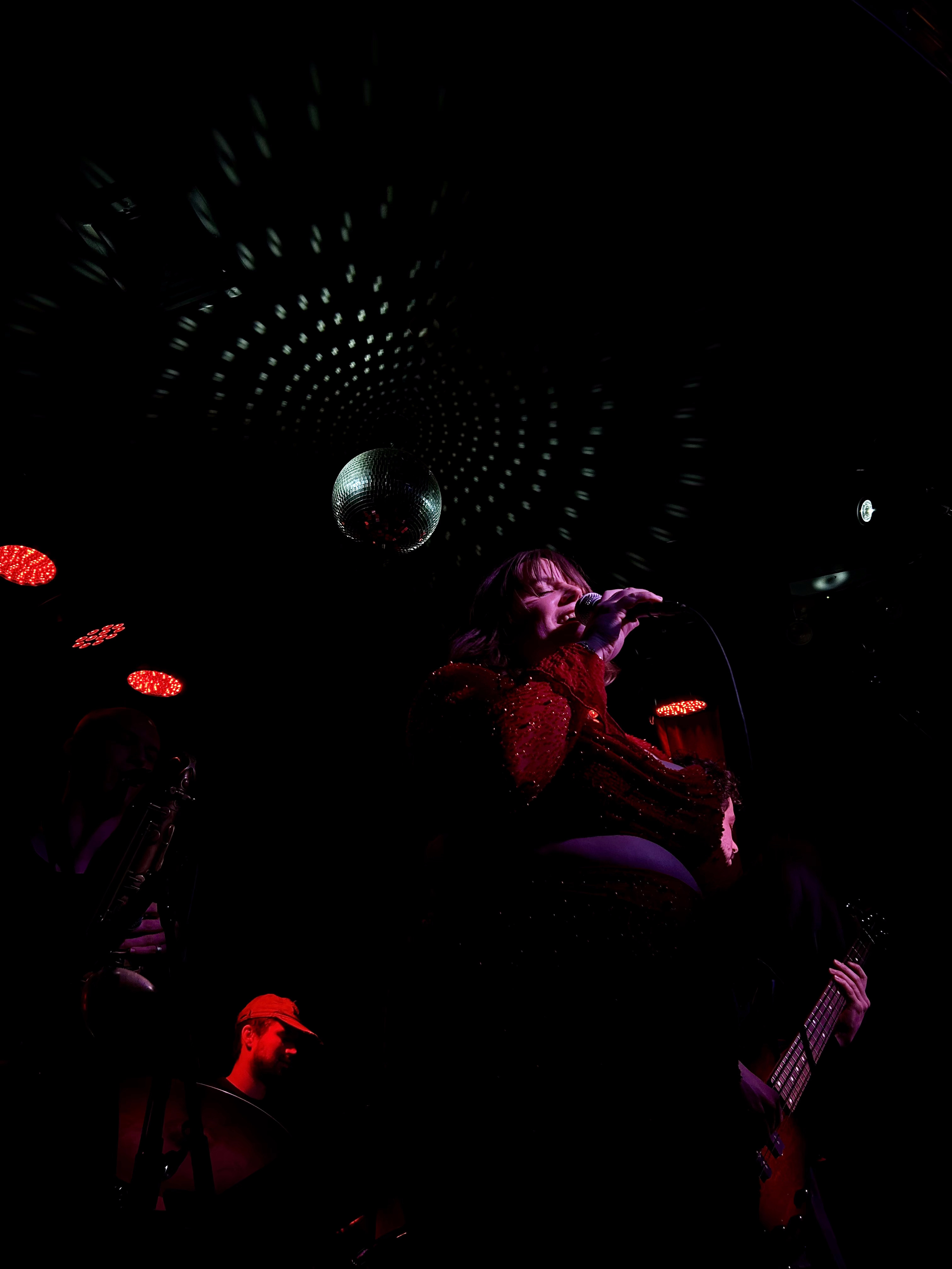 Taken from below, Moneypenny sings center, flanked by bandmates. The reflections of a discoball fills the ceiling above her.