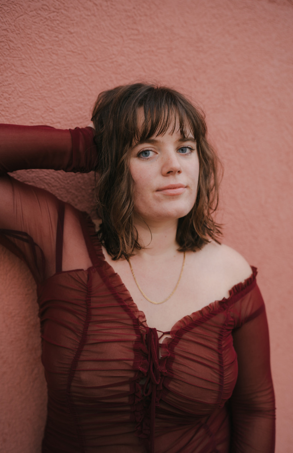 Madeline stands in a red gauze blouse, one hand behind her head, and leaning against a paler red, almost pink, wall.