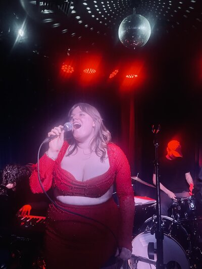 Madeline in a red dress, singing on stage beneath a disco ball.