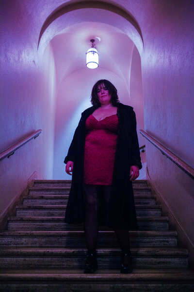 Madeline stands in a pastel purple stairwell looking down on the camera.